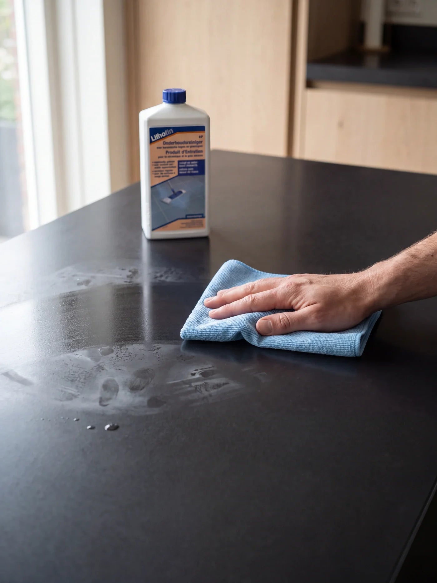Hand veegt een donker keramisch keukenblad schoon met een blauwe microvezeldoek, met Lithofin KF Onderhoudsreiniger ernaast